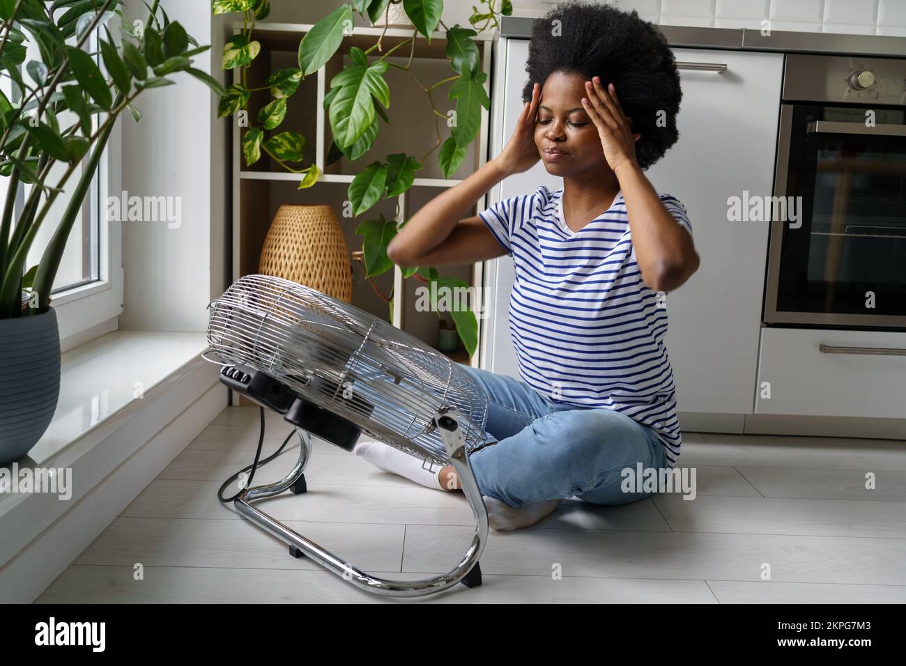 African woman suffering from heat at home, using electric fan for cooling during hot weather Stock Photo