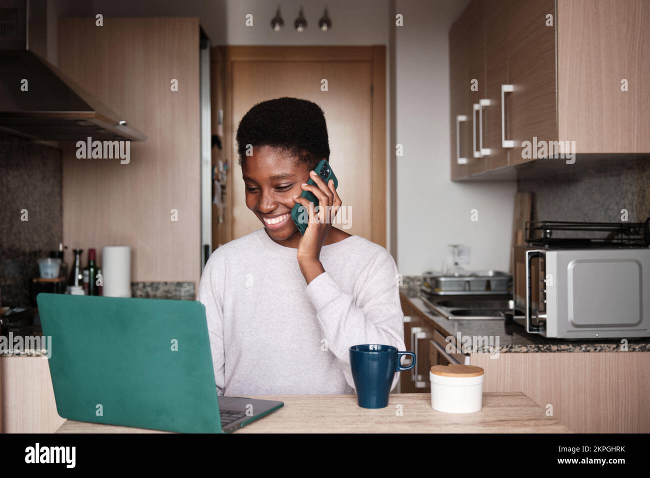 Smiling black woman talking on smartphone while using laptop Stock Photo