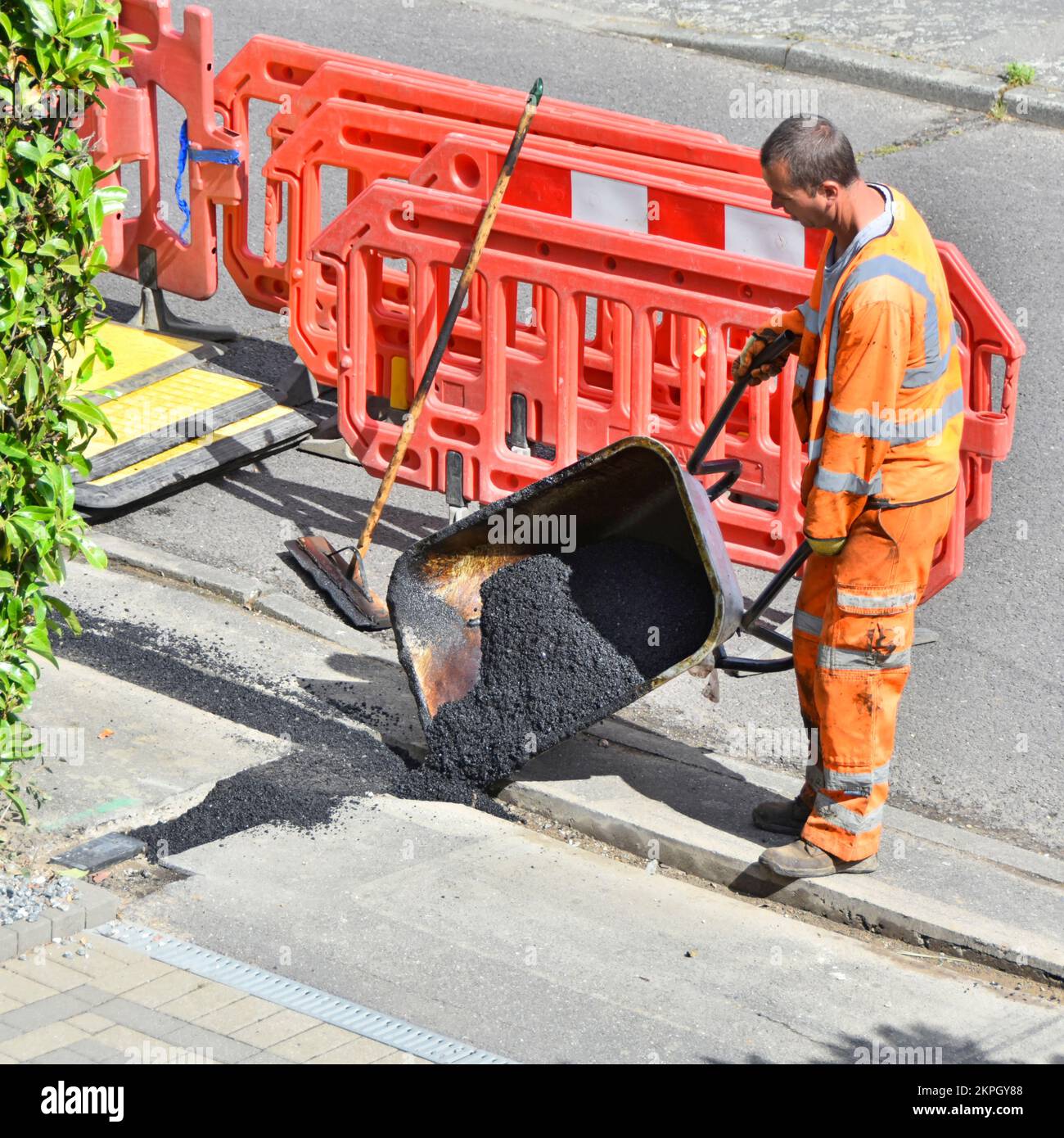 Road works workman wearing high visibility safety clothes tipping backfill tarmac out of wheelbarrow reinstating pavement broadband cable trench UK Stock Photo