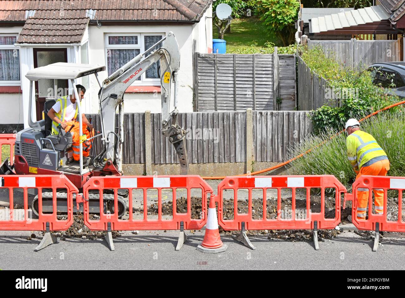 Mini excavator digger driver breaking up pavement for orange coloured fibre optic broadband cable in new trench assisted by labourer banksman Essex UK Stock Photo