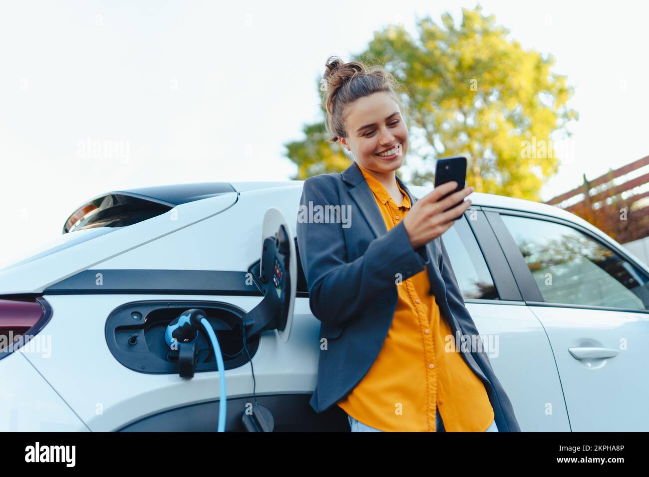Young woman with smartphone waiting while her electric car charging in home charging station, sustainable and economic transportation concept. Stock Photo