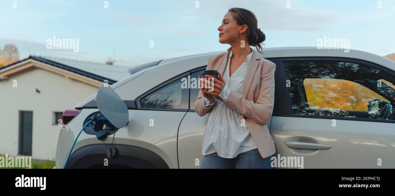 Young woman with cup of coffee waiting while her electric car charging, sustainable and economic transportation concept. Stock Photo