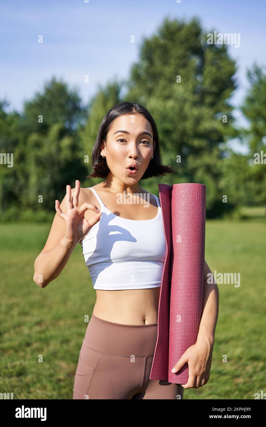Excited young woman standing with sports mat, yoga clothes, shows okay