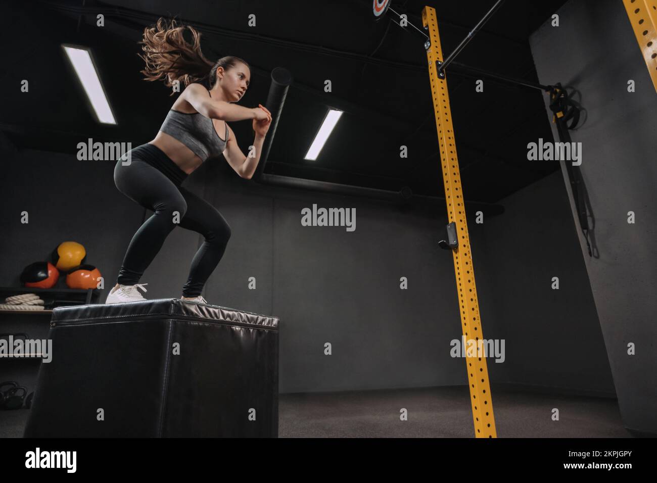 Woman doing box jump exercise as part of her crossfit training. Female athlete doing squats and jumping onto the box in dark workout gym. Copy space Stock Photo