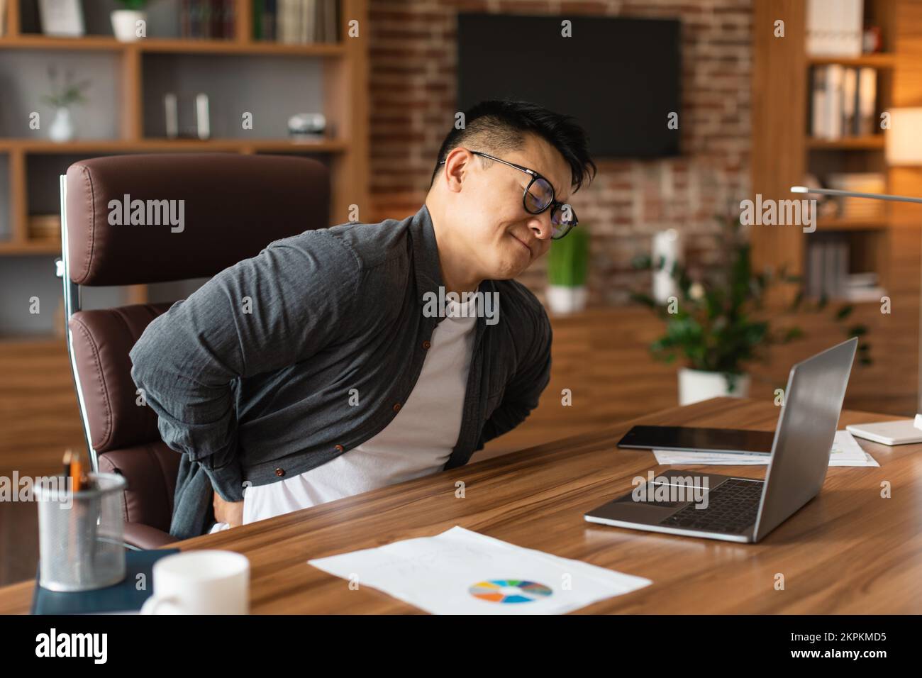 Unhappy middle aged asian man manager in glasses suffering from muscles back pain at table with laptop Stock Photo