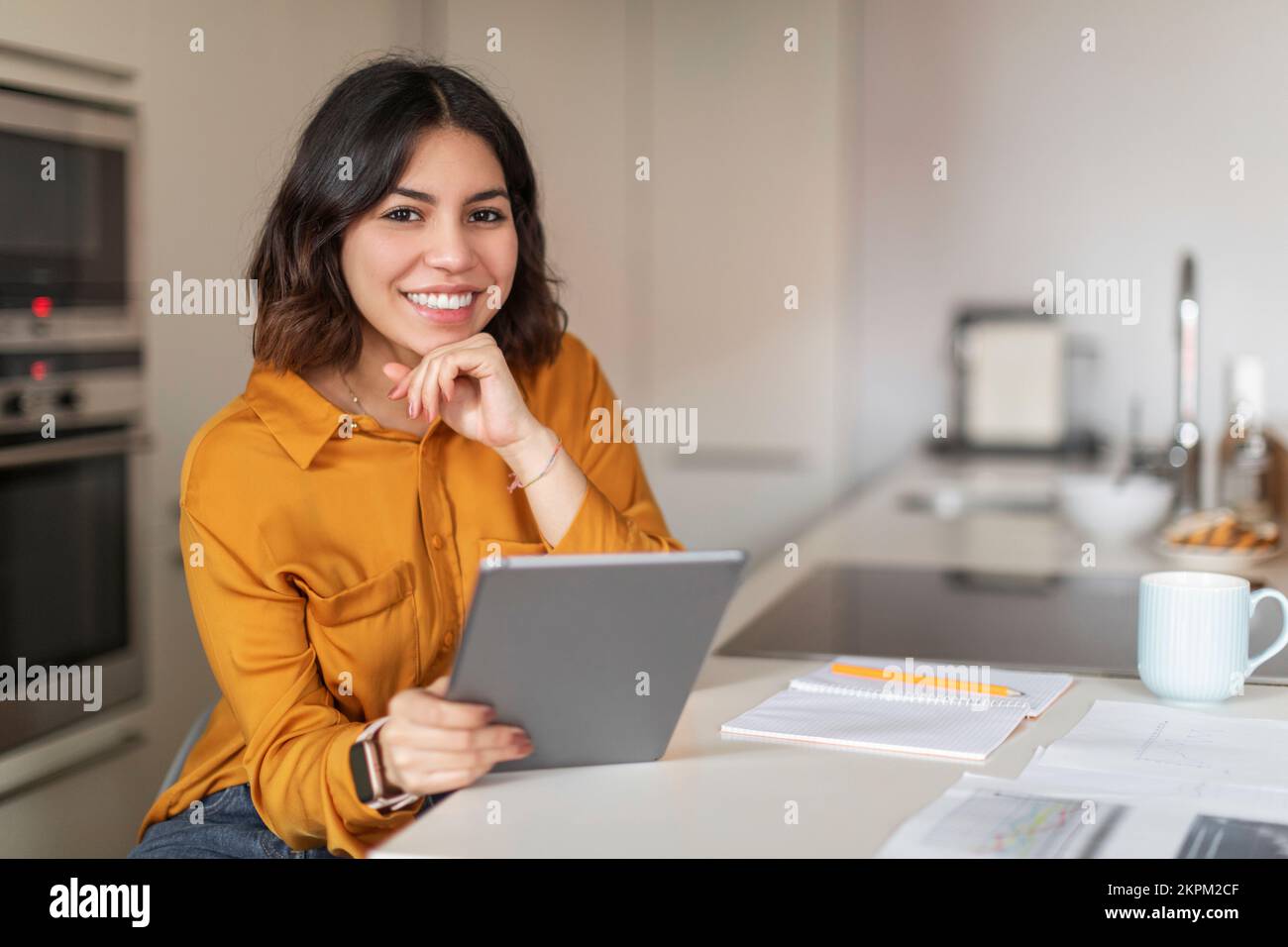 Smiling Young Arab Female Freelancer Using Digital Tablet While Sitting In Kitchen Stock Photo