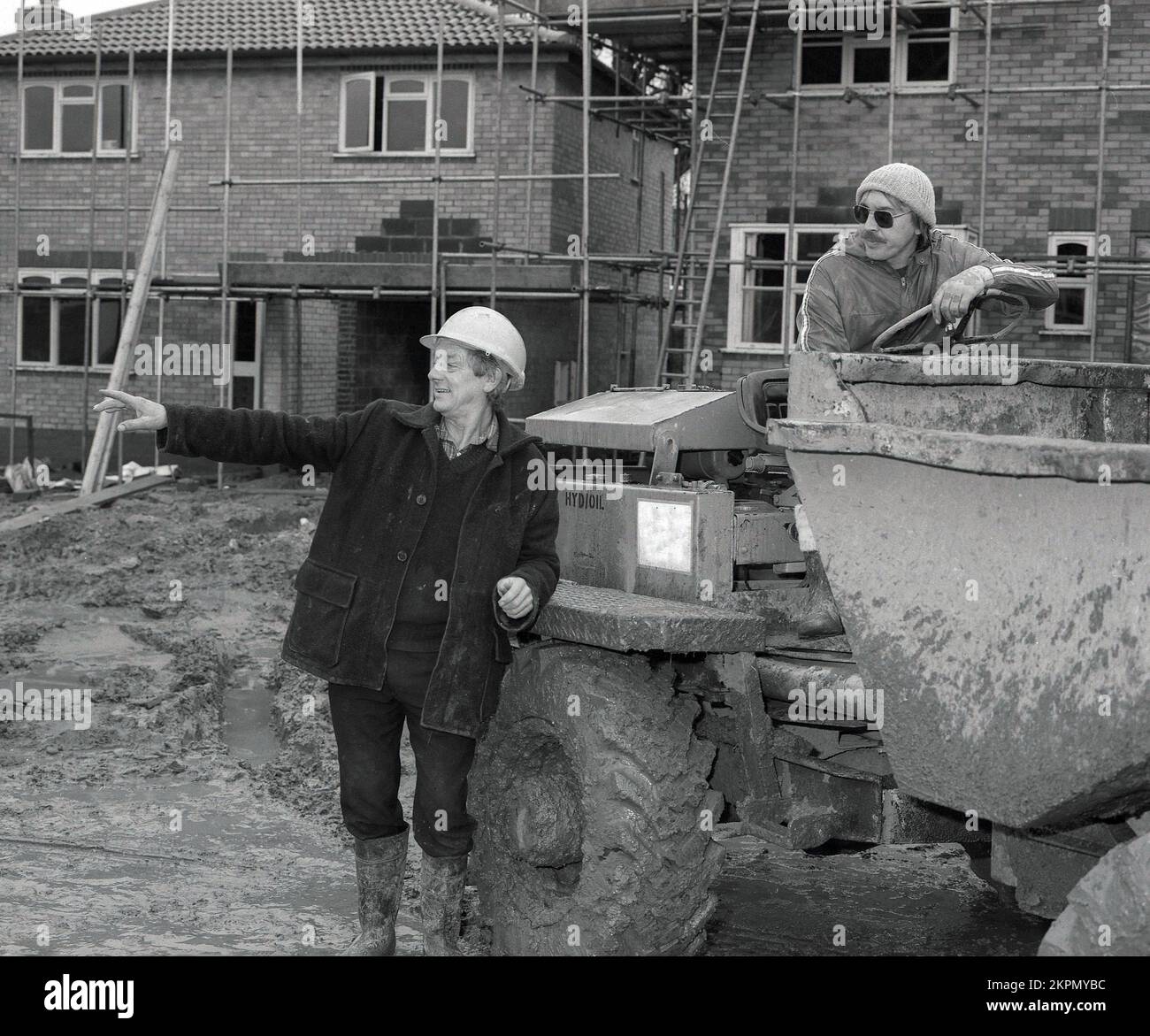 1980s, outside on a construction site of new housing, a foreman wearing