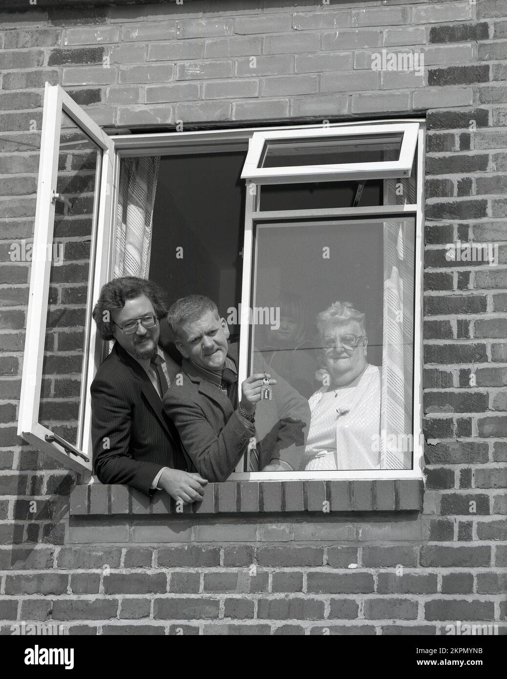 1980s, historical, standing at a window of their new home with a housing executive a couple with their daughter pose for a publicity photo, with the man holding the keys, England, UK. Stock Photo
