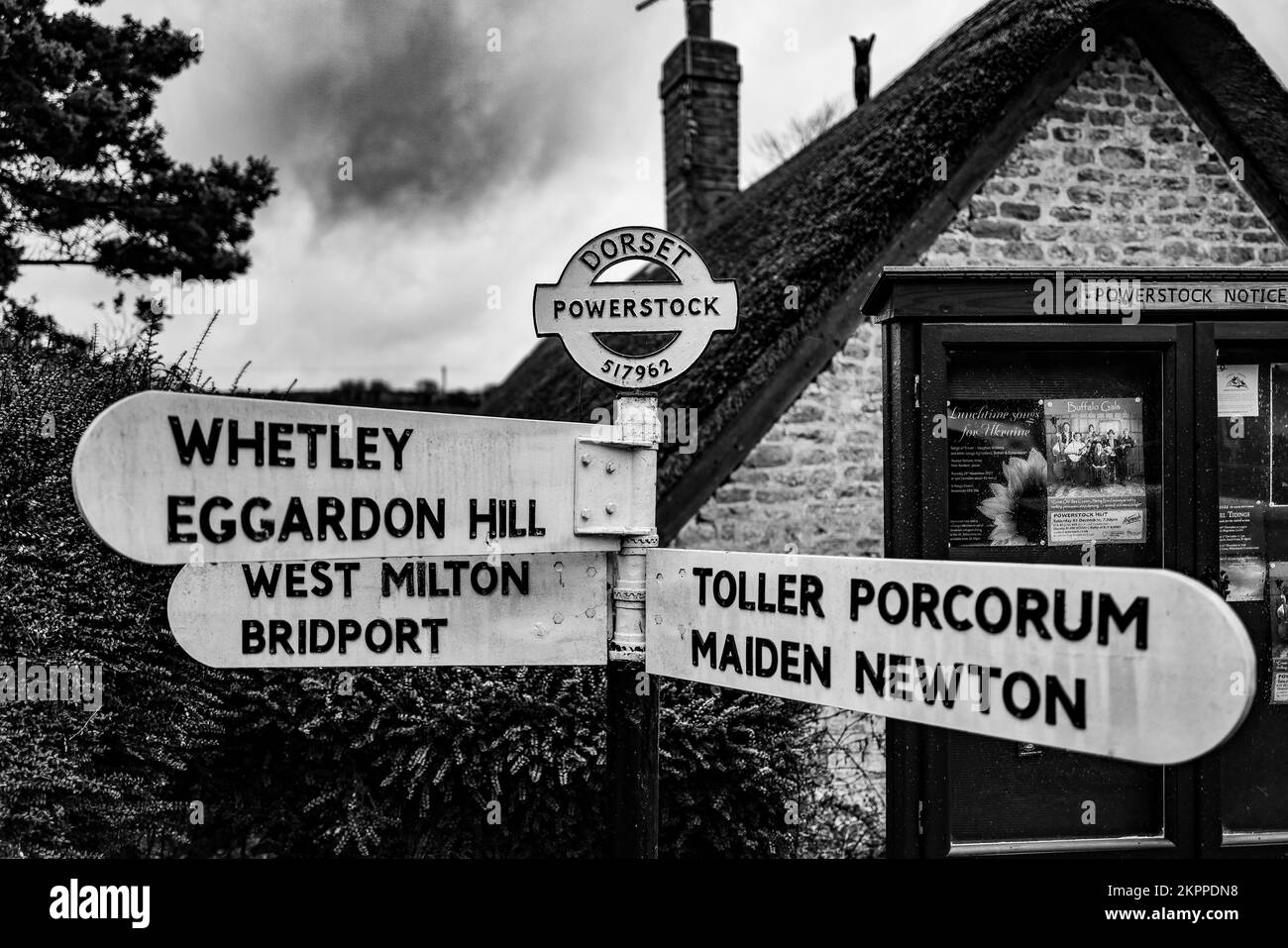 Public vintage road direction sign, in Dorset. Post standing in front of Powerstock Village notice board and cottage. B&W photo. Stock Photo