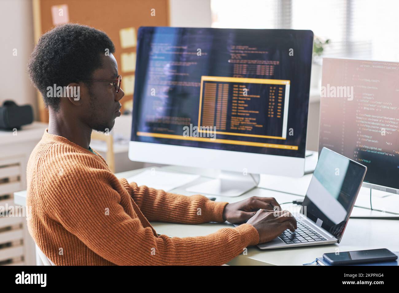 Serious young Black man in warm orange sweater concentrated on coding ...
