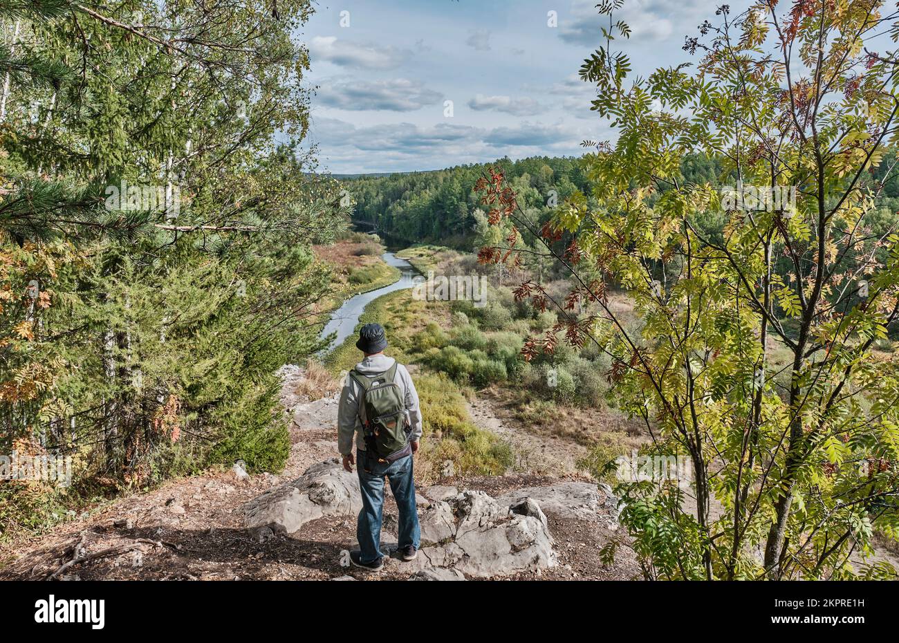 Back view of senior male hiker with backpack who is admiring landscape on top of rock. Nature park Deer streams, Sverdlovsk region, Russia. Serga Rive Stock Photo