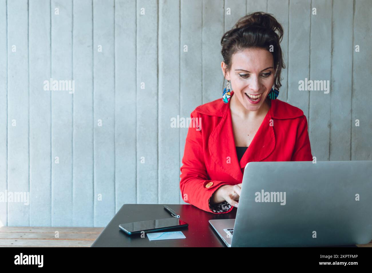 Adult business woman, dressed in red blazer, happy smiling seeing her business profit on the laptop, sitting working in a coffee shop. Stock Photo
