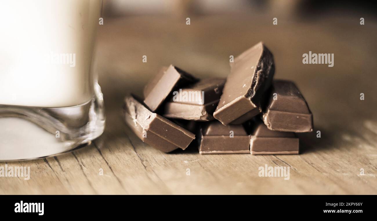 Shallow DOF macro photograph on a pile of sliced dark chocolate pieces sitting on wooden kitchen bench next to glass of milk Stock Photo