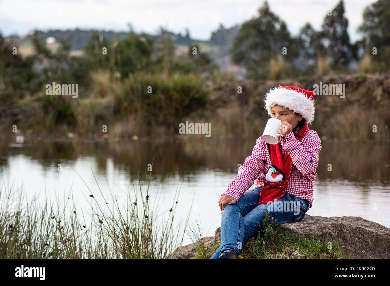 indigenous girl celebrating christmas with her christmas hat in the forest drinking coffee sitting on a stone Stock Photo