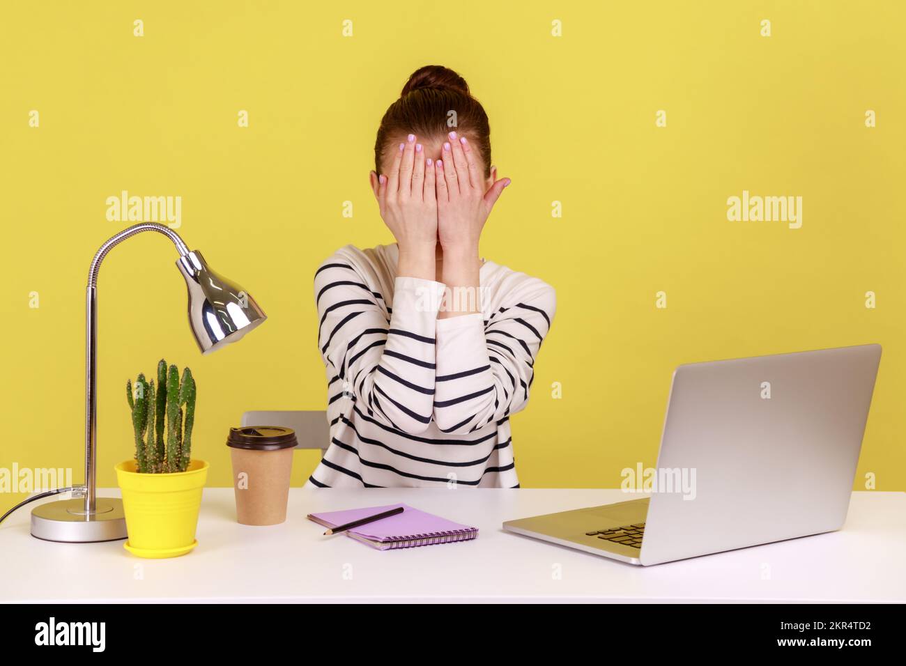 Don't want to see. Excited woman closing eyes with hand, avoid watching something shameful, working from home office. Indoor studio studio shot isolated on yellow background. Stock Photo