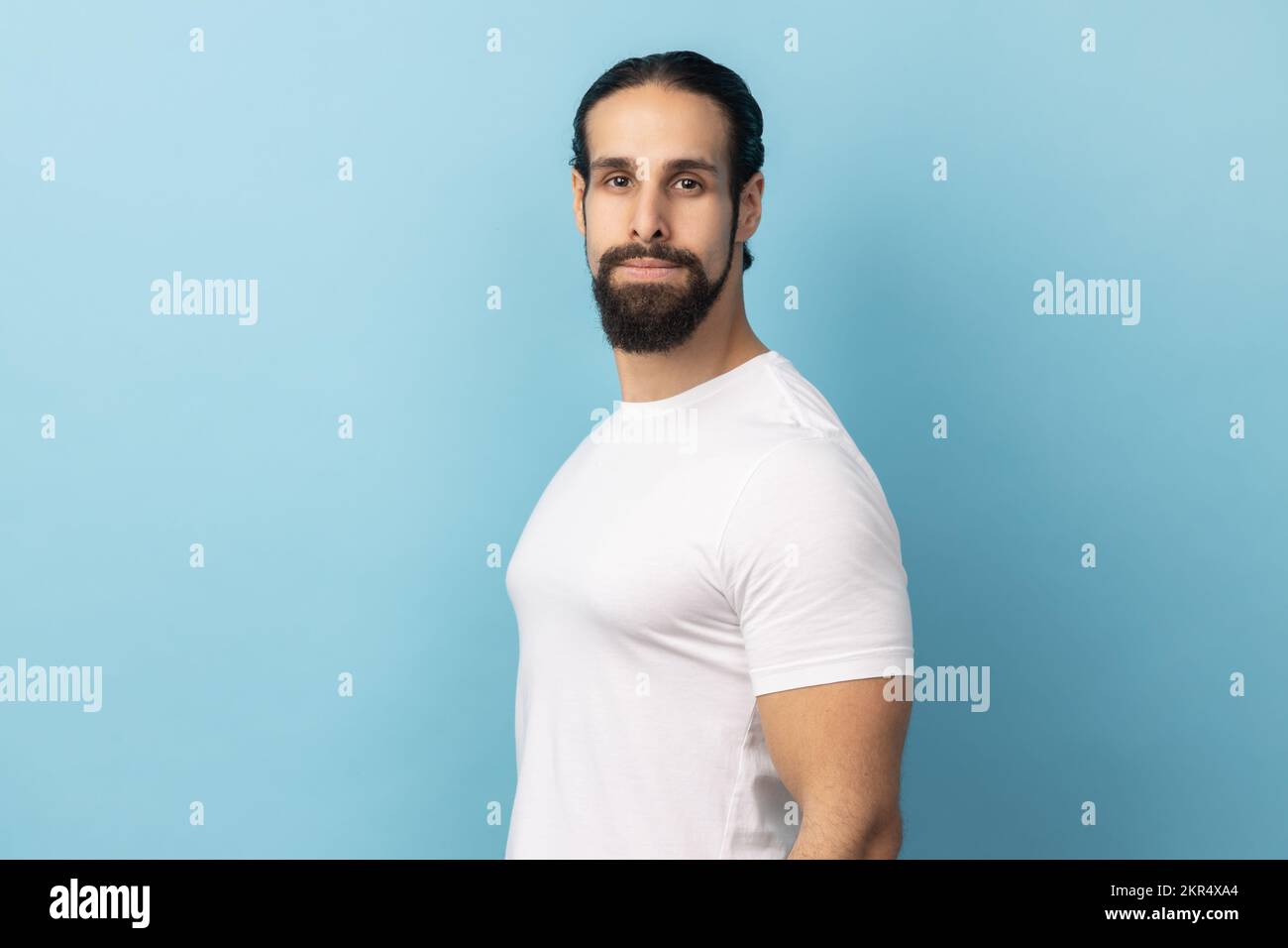Side view portrait of bearded handsome man wearing white T-shirt standing looking at camera with serious face, being strict and bossy. Indoor studio shot isolated on blue background. Stock Photo