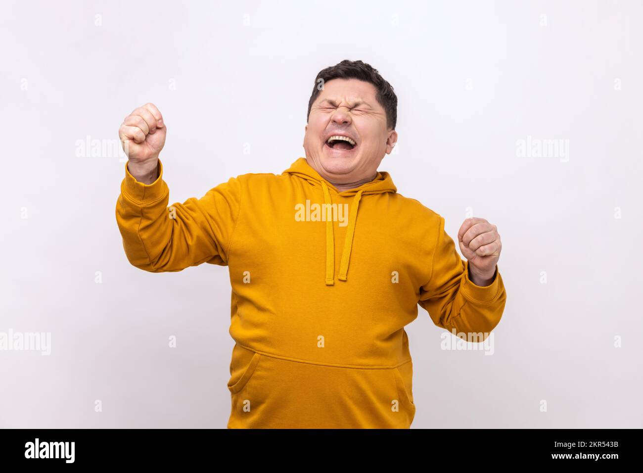 Overjoyed handsome middle aged man expressing winning gesture with raised fists and screaming, celebrating victory, wearing urban style hoodie. Indoor studio shot isolated on white background. Stock Photo
