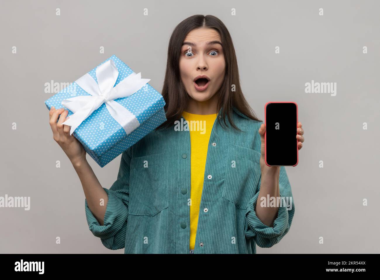 Astonished woman holding gift box and cell phone with empty display for online shopping advertising, expressing shock, wearing casual style jacket. Indoor studio shot isolated on gray background. Stock Photo