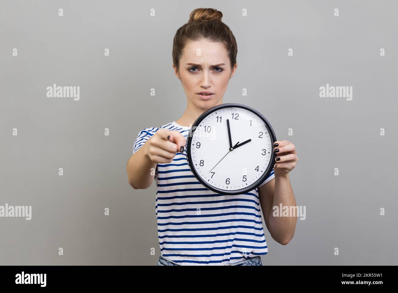 Portrait of bossy serious woman wearing striped T-shirt pointing finger at you holding in hand big wall clock, motivation. Indoor studio shot isolated on gray background. Stock Photo