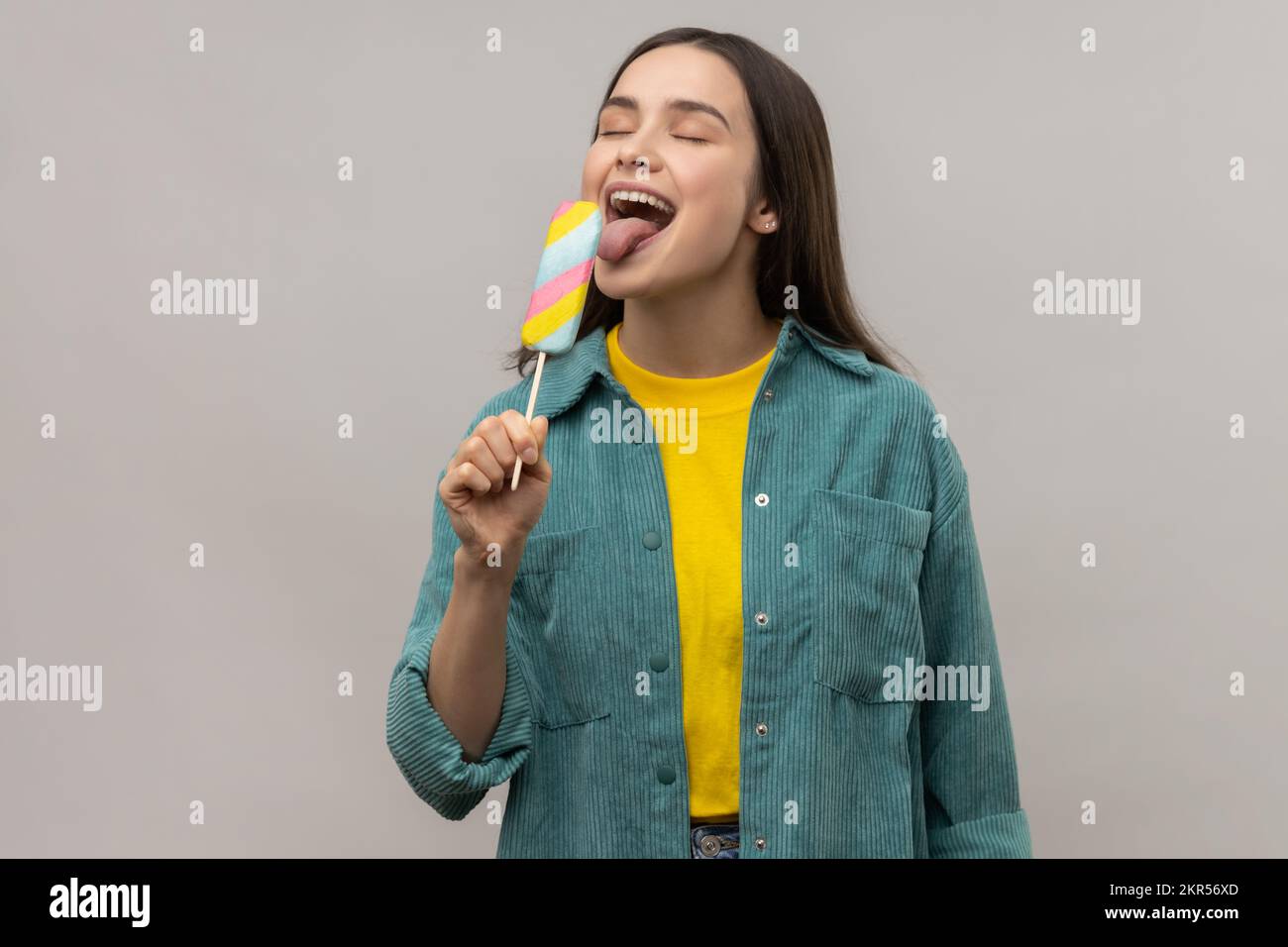 Portrait of positive woman with dark hair licking multicolored ice cream, feels hungry, tasting dessert, wearing casual style jacket. Indoor studio shot isolated on gray background. Stock Photo