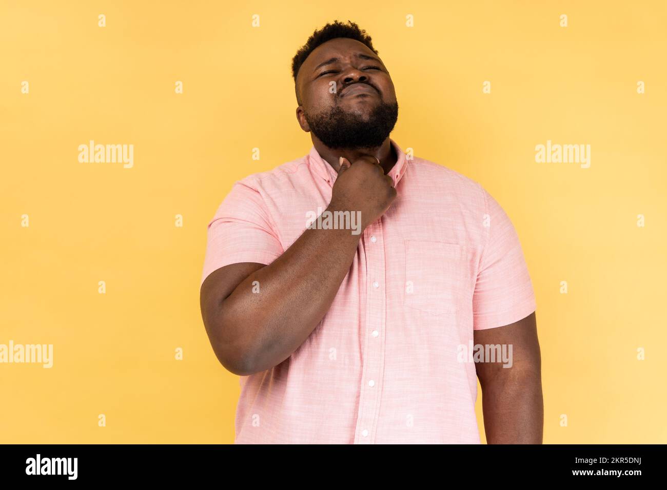Throat pain. Portrait of sick man wearing pink shirt standing and touching his painful throat, frowning his face, feels terrible pain. Indoor studio shot isolated on yellow background. Stock Photo