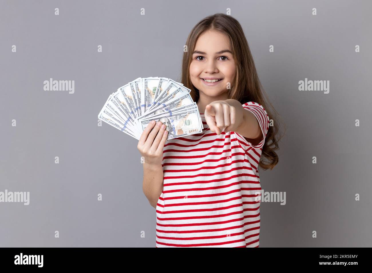 Portrait of delighted little girl wearing striped showing at fan of dollar banknotes, showing big money and pointing at camera. T-shirt Indoor studio shot isolated on gray background. Stock Photo