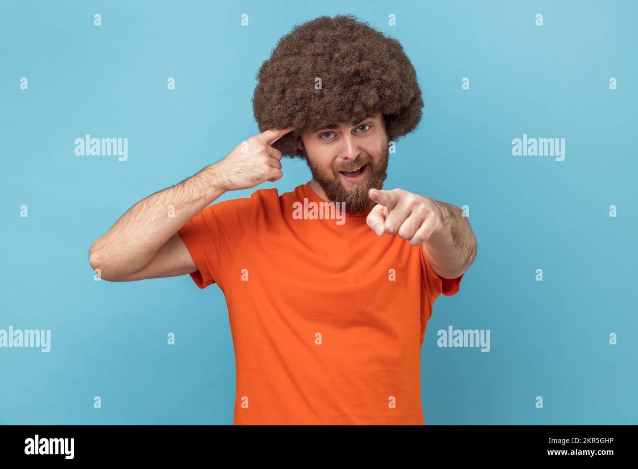 Hey, you are kidding. Man with Afro hairstyle surprised face holding finger near head and pointing finger at camera, stupid gesture. Indoor studio shot isolated on blue background. Stock Photo