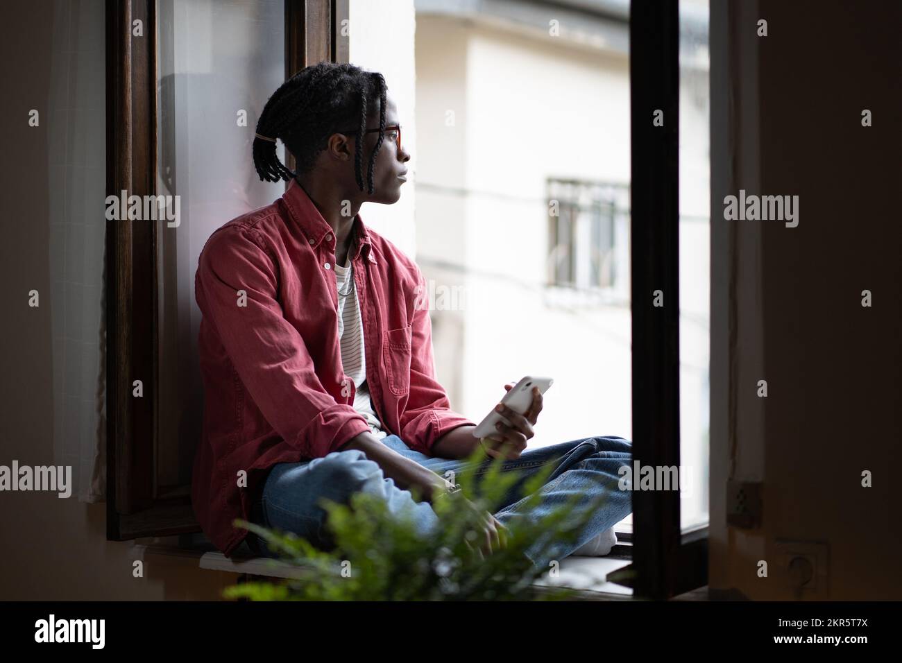 African student guy sitting on windowsill holding smartphone, missing friends while studying abroad Stock Photo