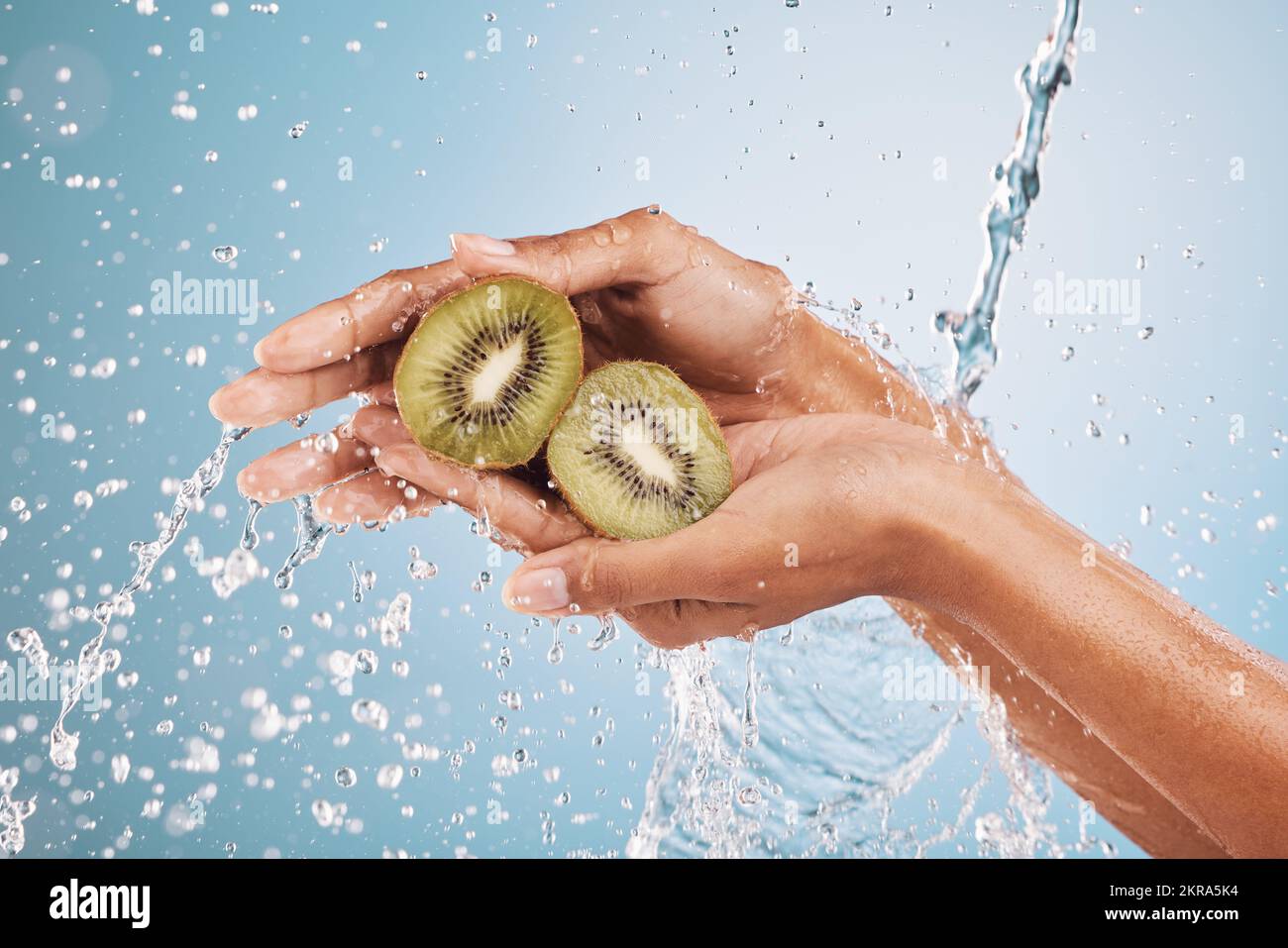 Water splash, hands of woman and kiwi in studio on a blue background ...