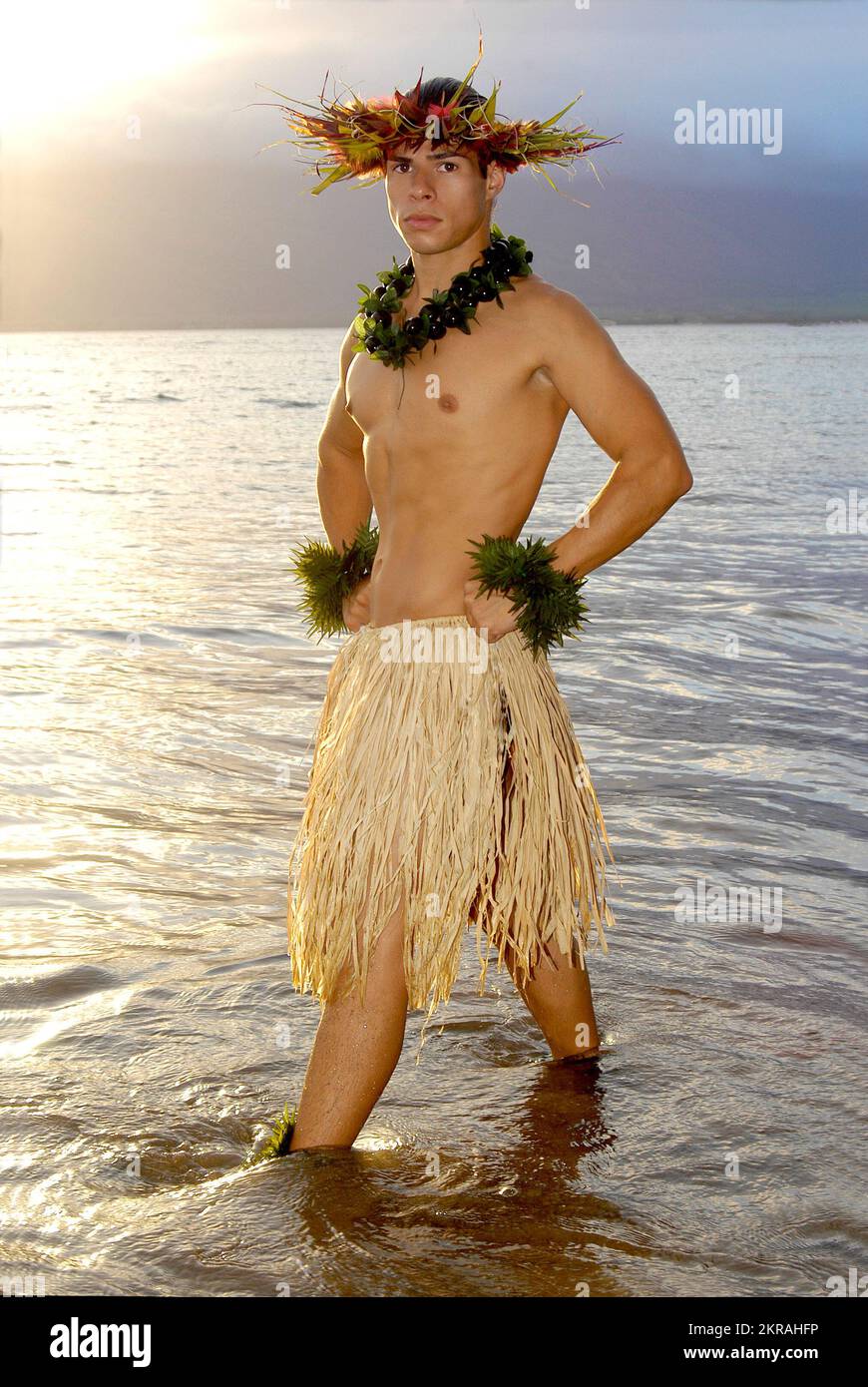 Handsome Male Hula Dancer stands in the shallow part of the ocean as the sun sets behind him. Stock Photo