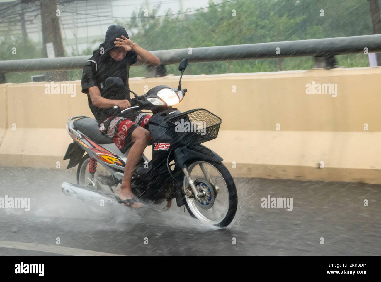 Ratchaburi, Thailand, NOV 14 2022, A young man is riding a motorcycle, shielding his eyes from the heavy rain with his hand Stock Photo