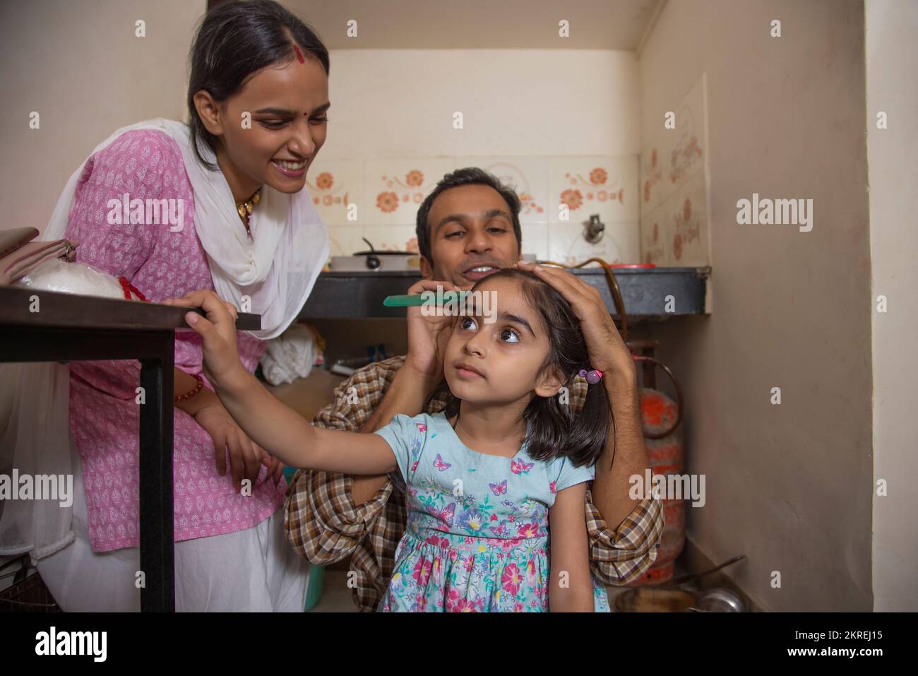 Father combing his daughter's hair in the kitchen while mother standing beside Stock Photo