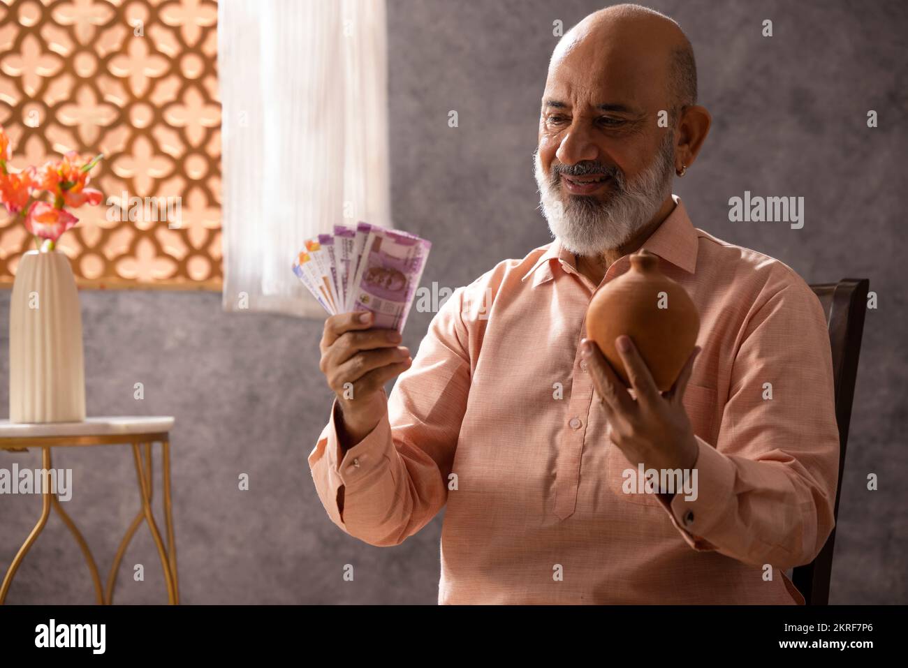Portrait of happy senior man sitting at home with piggy bank and Indian ...
