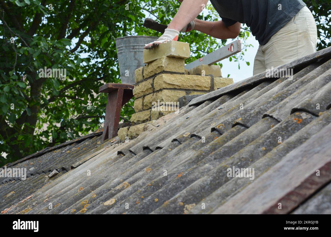 Chimney repair with laying new bricks. Building contractor is bricklaying new chimney pipe on old house with asbestos roof. Stock Photo