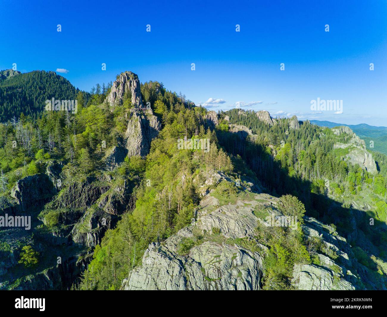 Rocky peak of high mountain with vegetation in valley of Rhodope mountains and forests against background of sky with large clouds Stock Photo