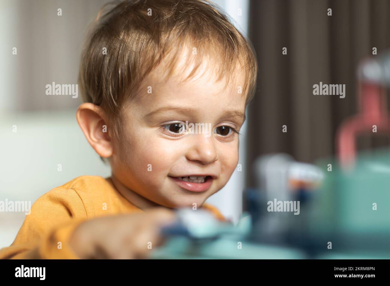 Cheerful adorable little blond haired boy in orange sweater sitting at table and playing with toy while spending time at home Stock Photo