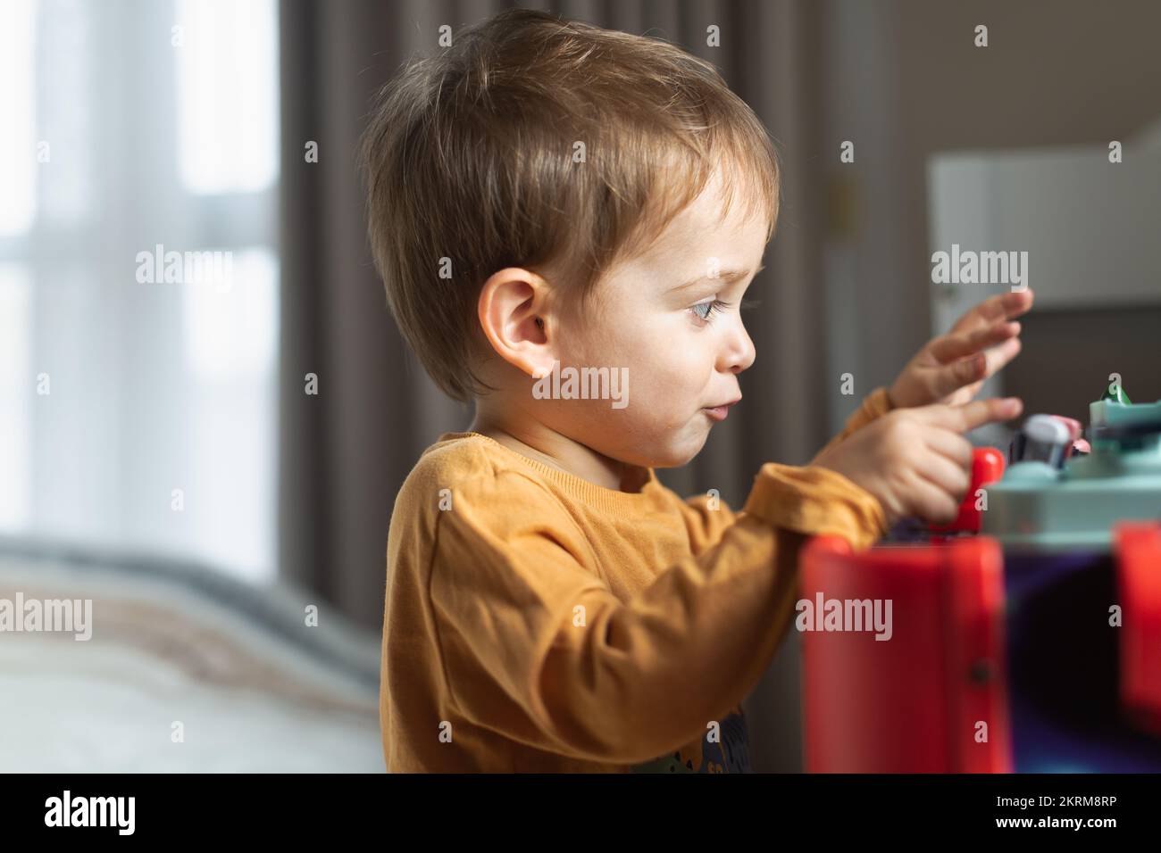 Side view of adorable little blond haired boy in orange sweater sitting at table and playing with toy while spending time at home Stock Photo