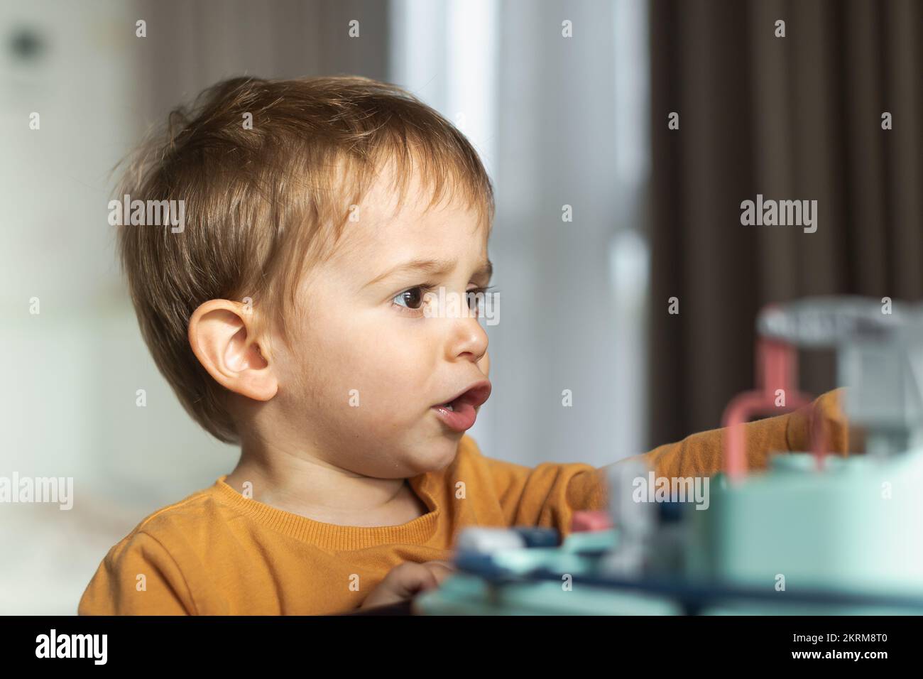 Adorable little blond haired boy in orange sweater sitting at table and playing with toy while spending time at home Stock Photo