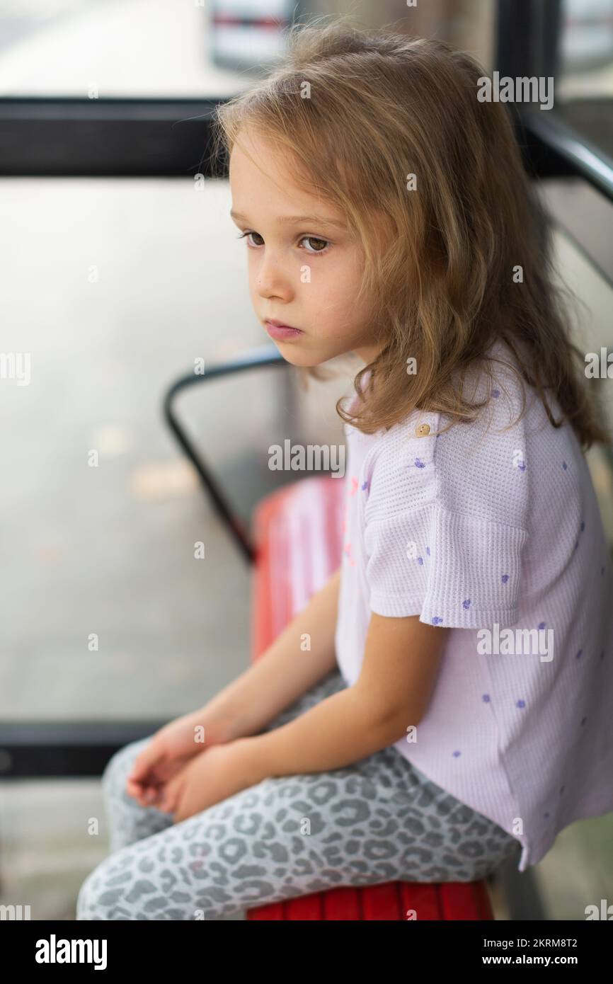 High angle of calm little girl with blond hair in purple t shirt and gray leopard leggings sitting at bus stop Stock Photo