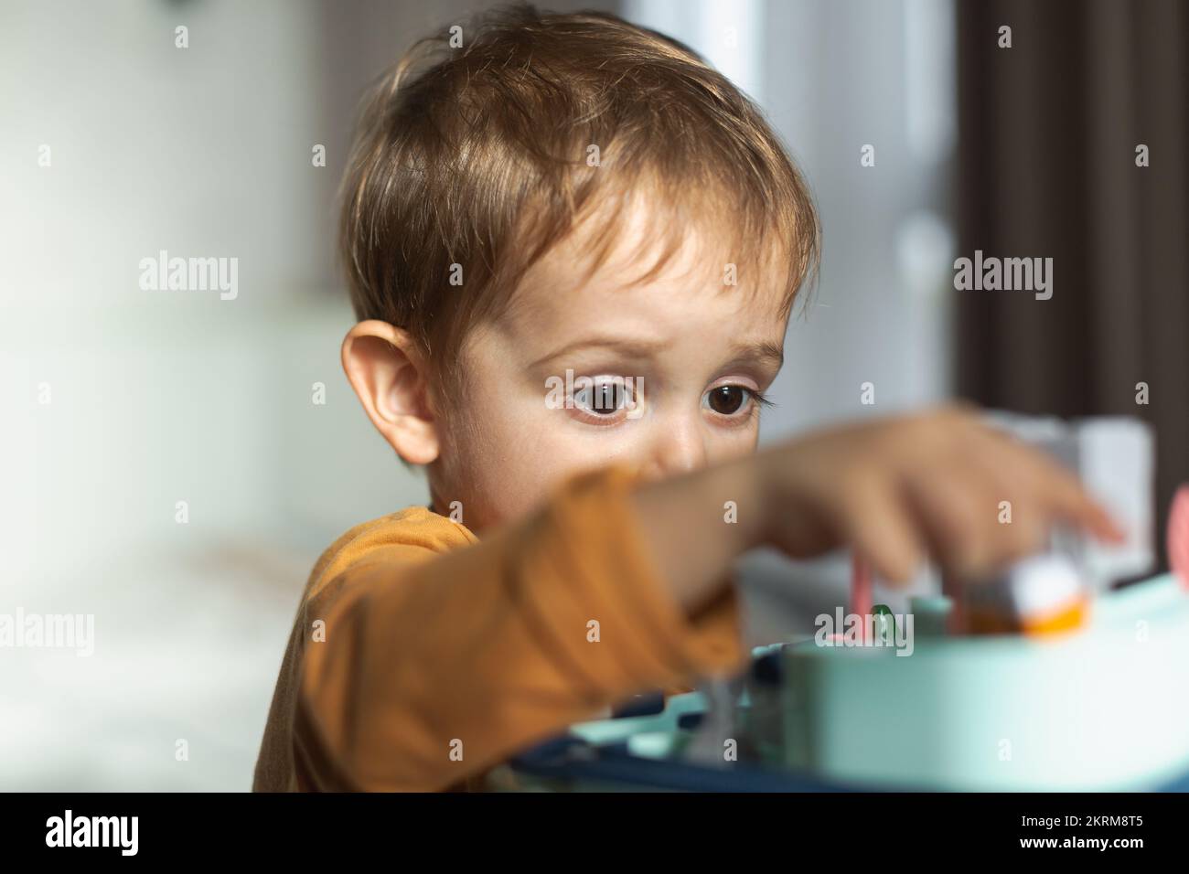 Adorable little blond haired boy in orange sweater sitting at table and playing with toy while spending time at home Stock Photo