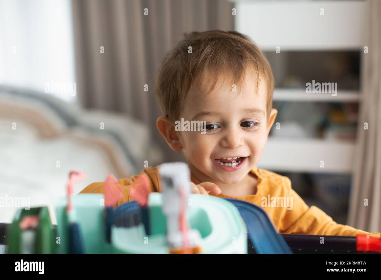 Cheerful adorable little blond haired boy in orange sweater sitting at table and playing with toy while spending time at home Stock Photo