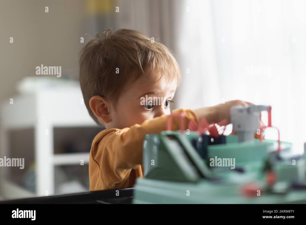 Side view of adorable little blond haired boy in orange sweater sitting at table and playing with toy while spending time at home Stock Photo