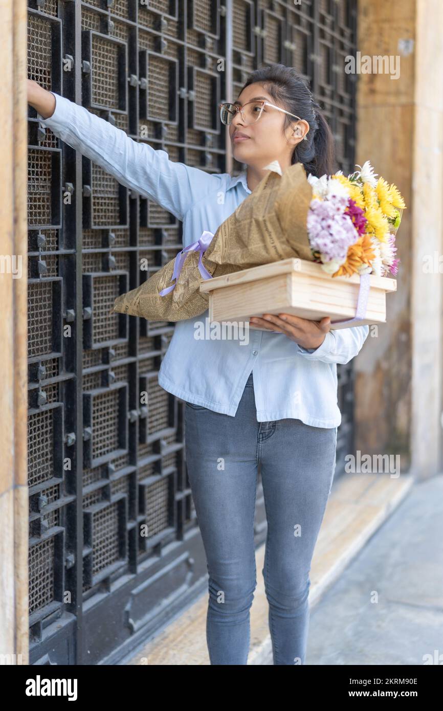 Positive woman in casual apparel and eyeglasses standing near metal door with bouquet of colorful flowers and wooden gift box ringing doorbell Stock Photo