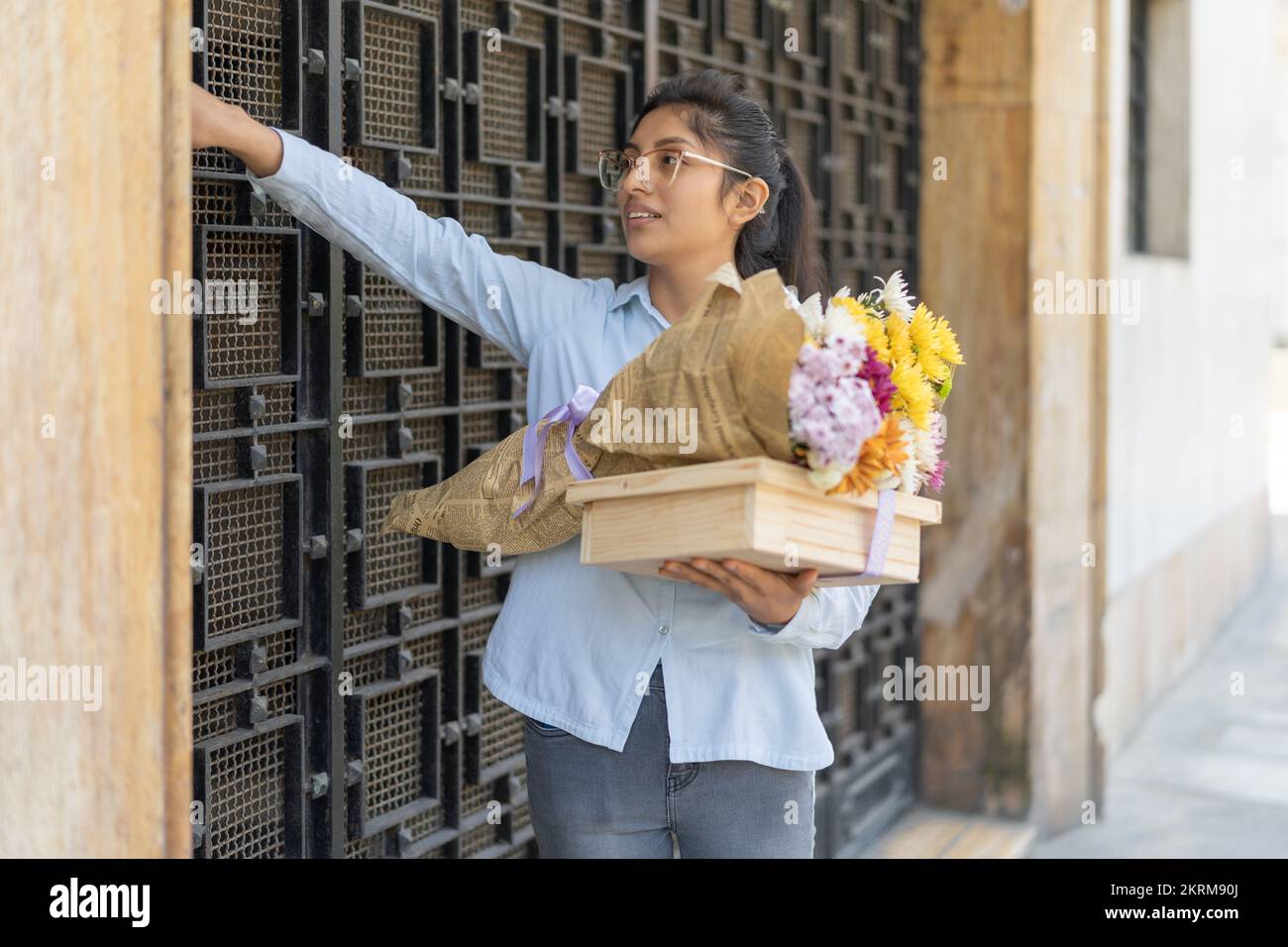 Positive woman in casual apparel and eyeglasses standing near metal door with bouquet of colorful flowers and wooden gift box ringing doorbell Stock Photo