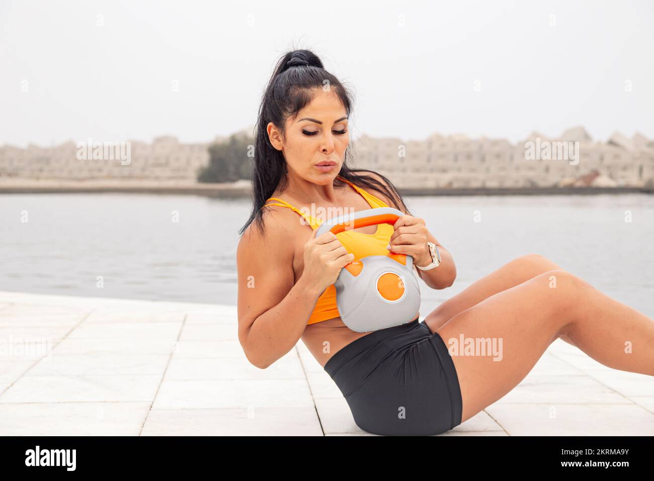 Fit female athlete in tight sportswear sitting on tiles while doing exercises with kettlebell during fitness workout near water Stock Photo