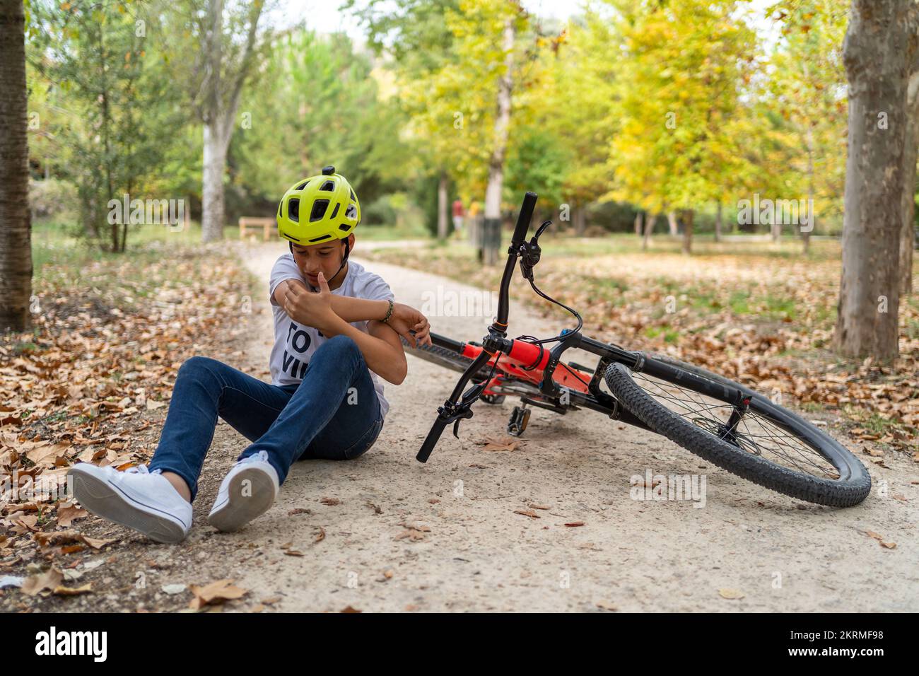 Full body of boy in helmet sitting on ground pathway near red bicycle