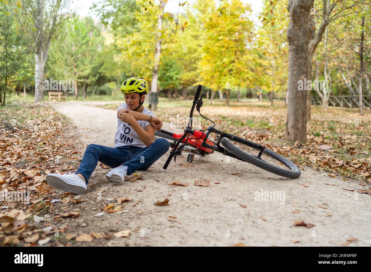 Full body of boy in helmet sitting on ground pathway near red bicycle