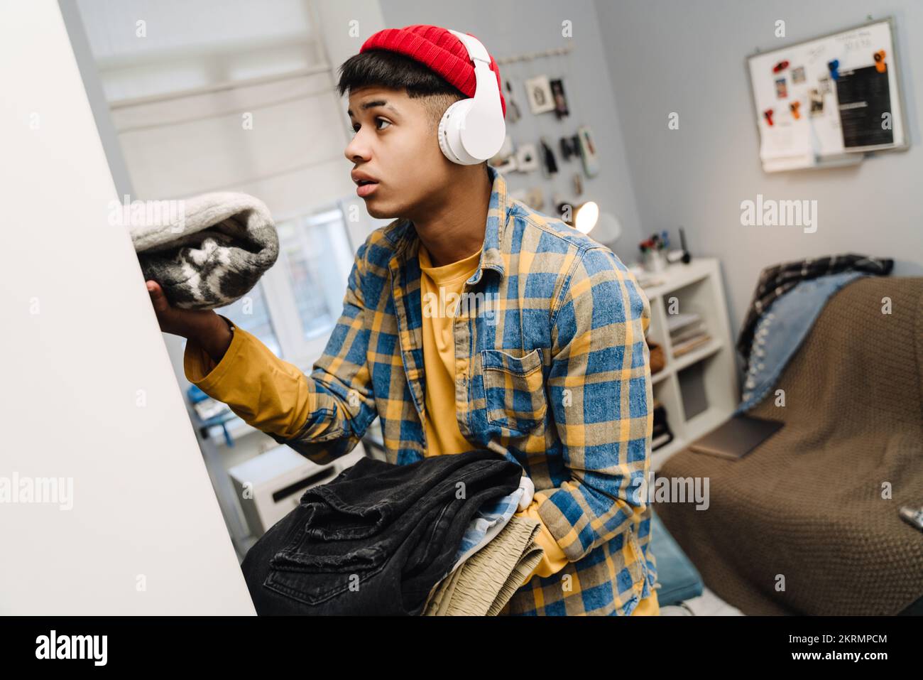 Teenage boy using headphones folding his clothes while cleaning bedroom at home Stock Photo