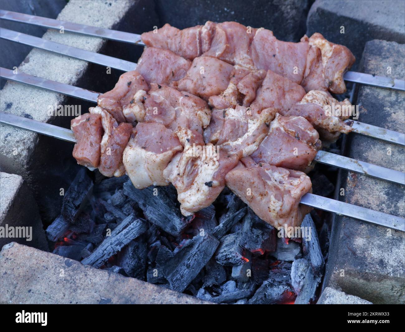 raw meat on four skewers grilled on brick-lined coals, classic pork skewers in the first stage of cooking on a smoking fire on a camping trip Stock Photo