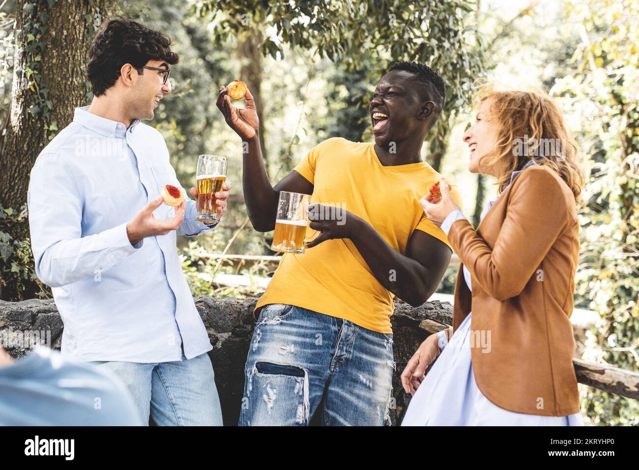 multiracial group of friends laughing and having fun at party, mixed age range group celebrating something eating snack and drinking beers Stock Photo
